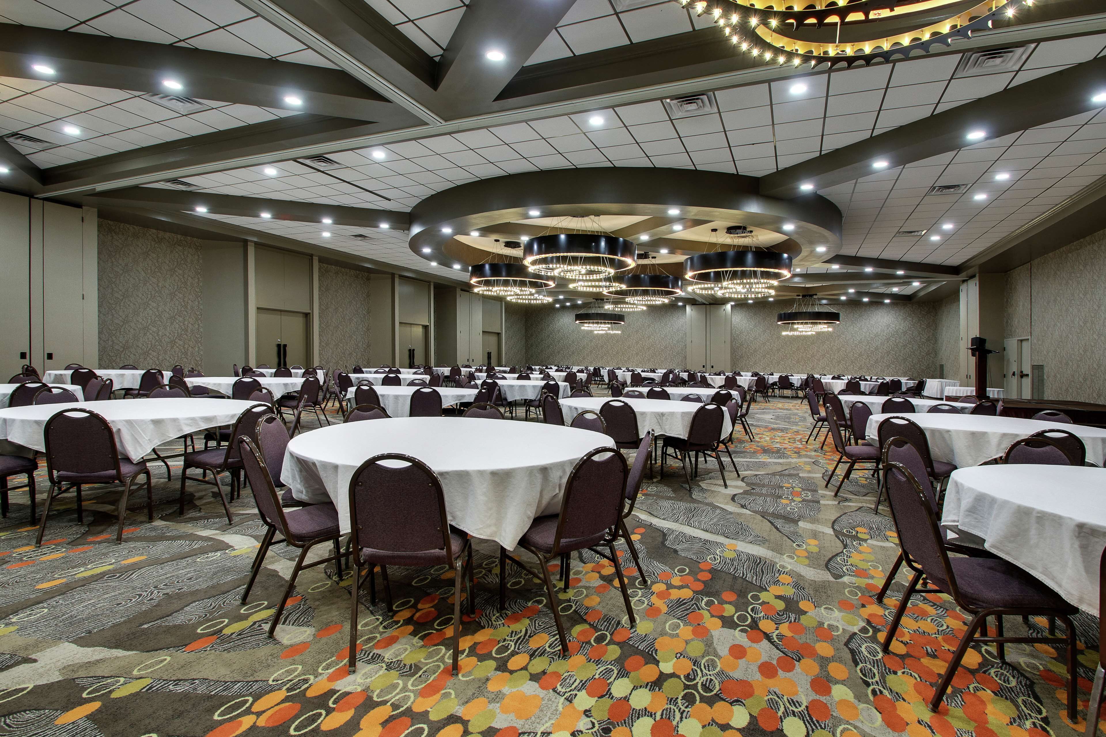 A large banquet hall with round tables and chairs, a patterned carpet, and a modern chandelier.