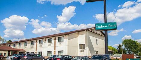 A hotel with a sign indicating an indoor pool, a parking lot with several cars, and a clear blue sky with clouds.