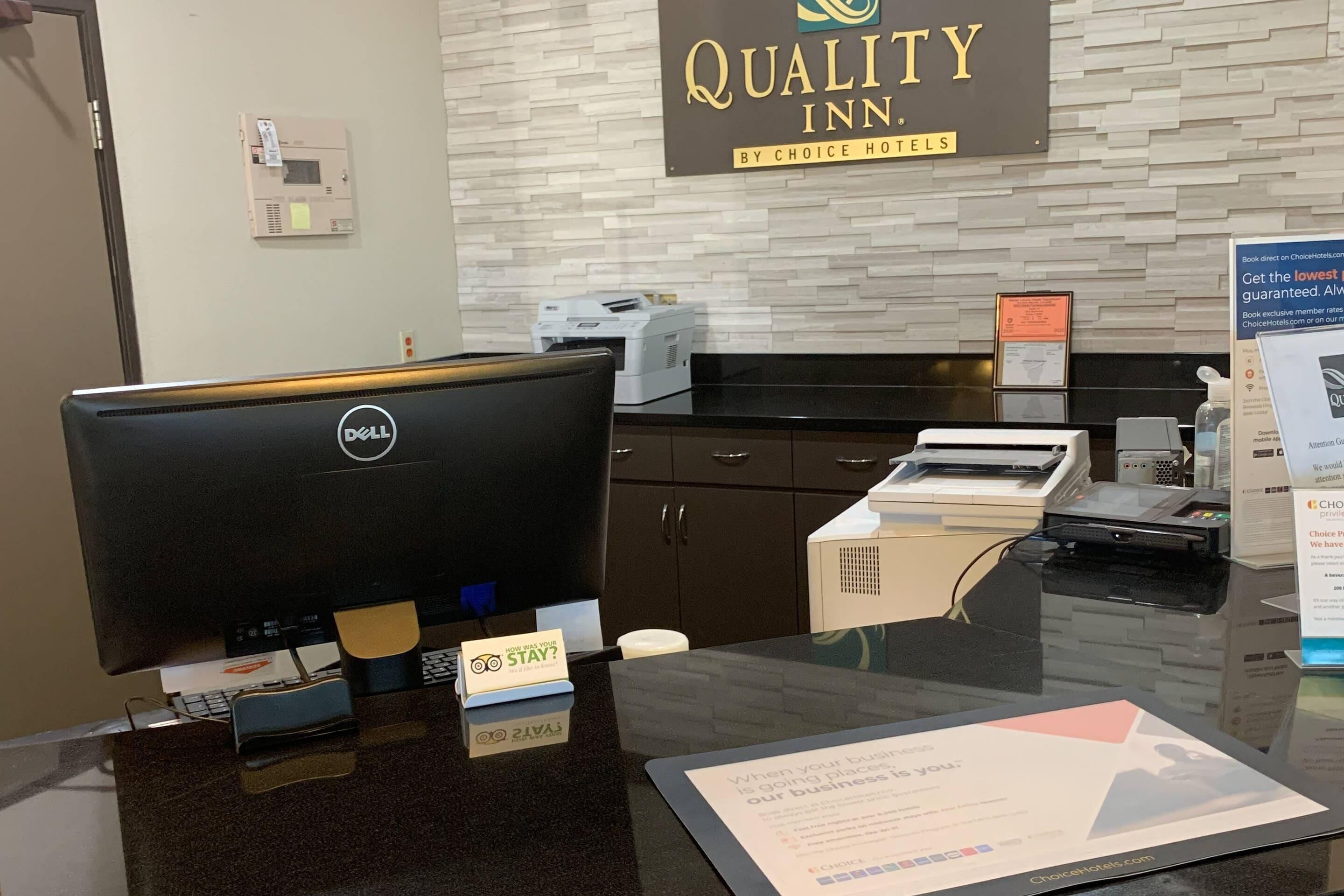 Un comptoir d’accueil d’hôtel avec un ordinateur, une enseigne « Quality Inn » et une affiche imprimée accrochée au mur.