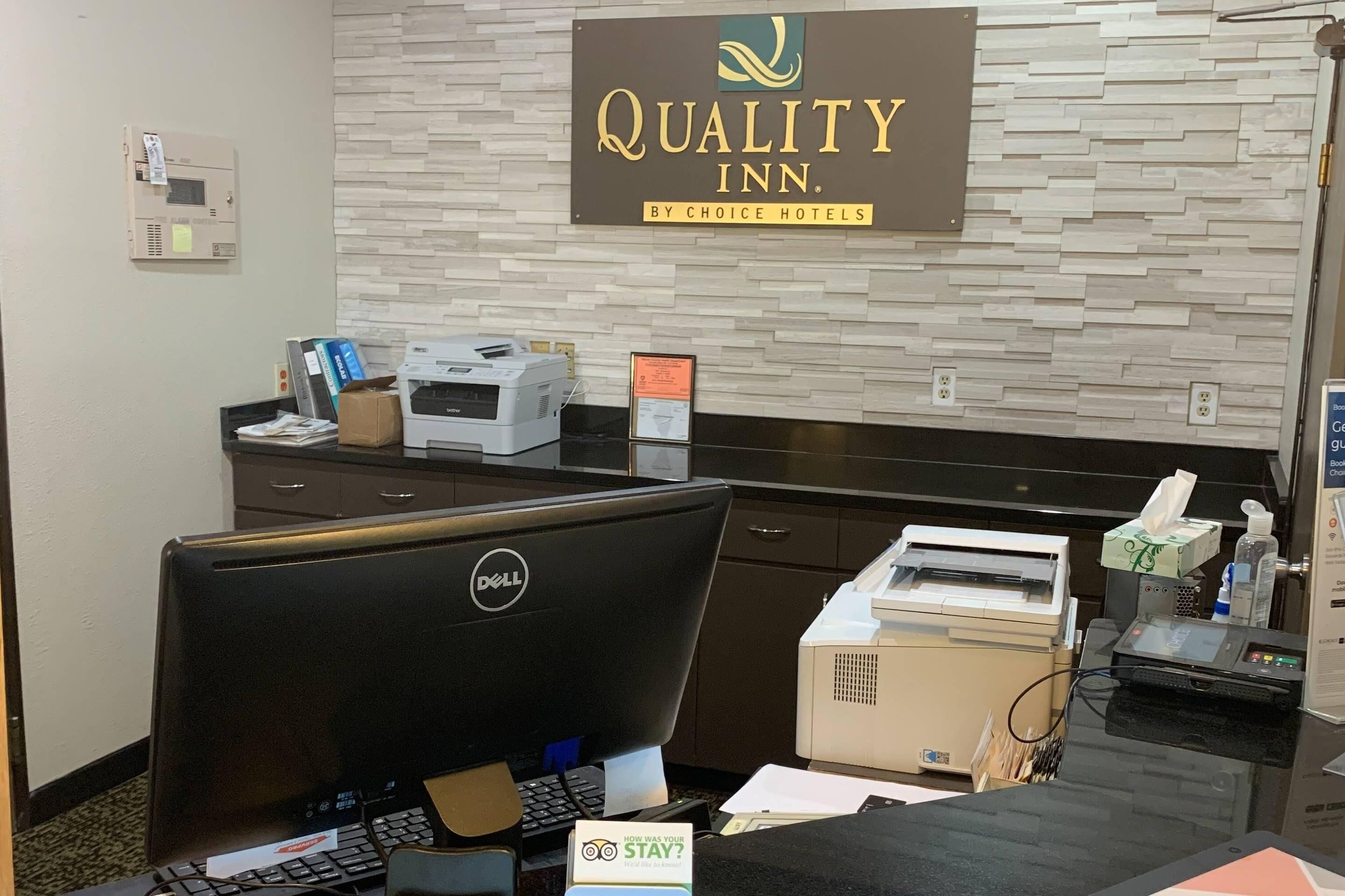 A hotel reception desk with a sign for Quality Inn by Choice Hotels, a computer, printer, and various office supplies.