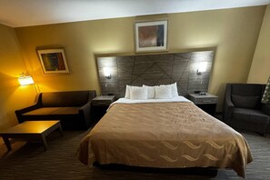 A hotel room with a large bed, a sofa, a small table, two nightstands, and a wall-mounted lamp. - Quality Inn Forsyth - Decatur (Forsyth)