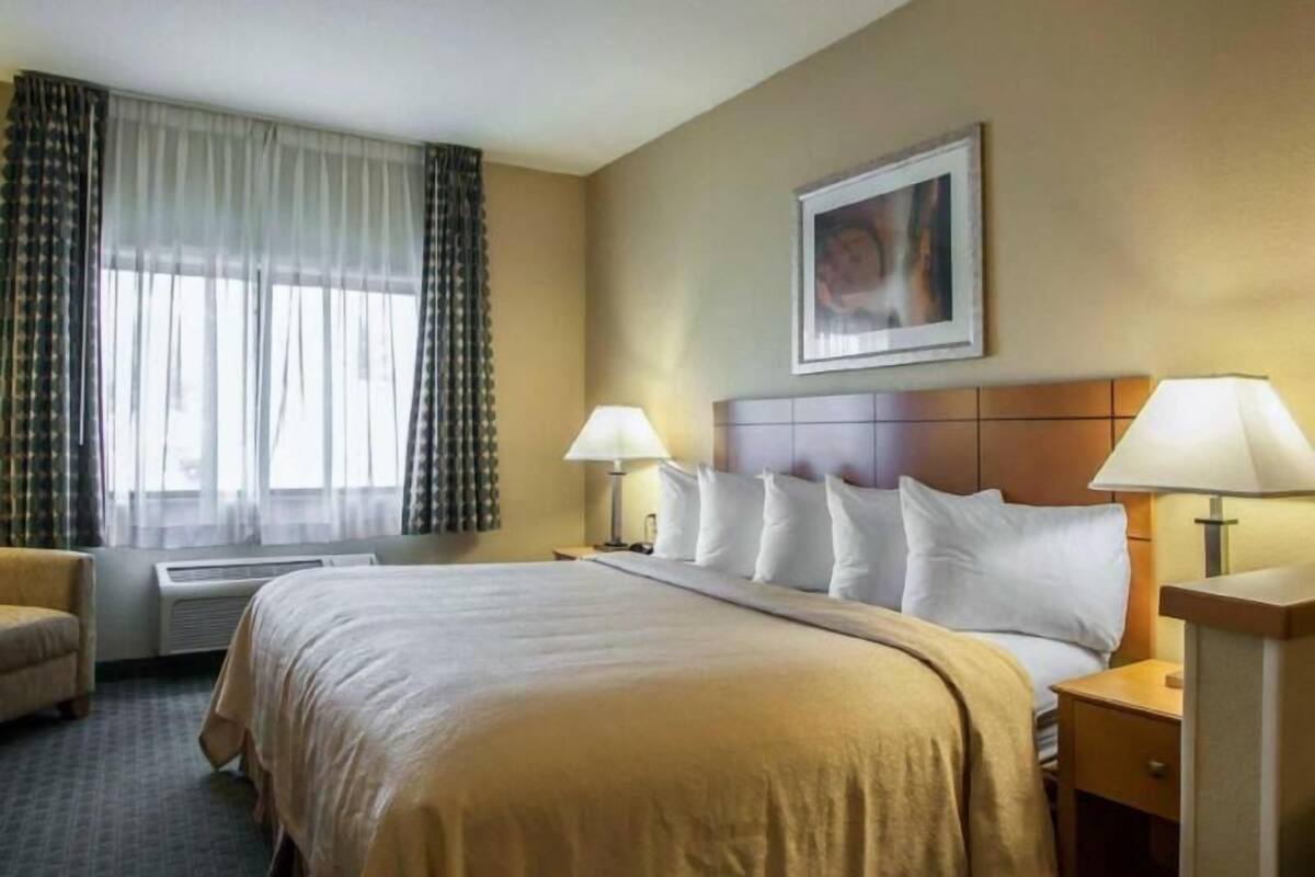 Una habitación de hotel con cama, dos lámparas de noche, cabecera, un cuadro enmarcado en la pared y una ventana con cortinas.