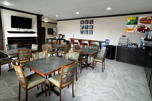 A dining area with multiple tables and chairs, a flat-screen TV mounted above a fireplace, and a kitchen area with a coffee station. - Quality Inn Forsyth - Decatur (Forsyth)