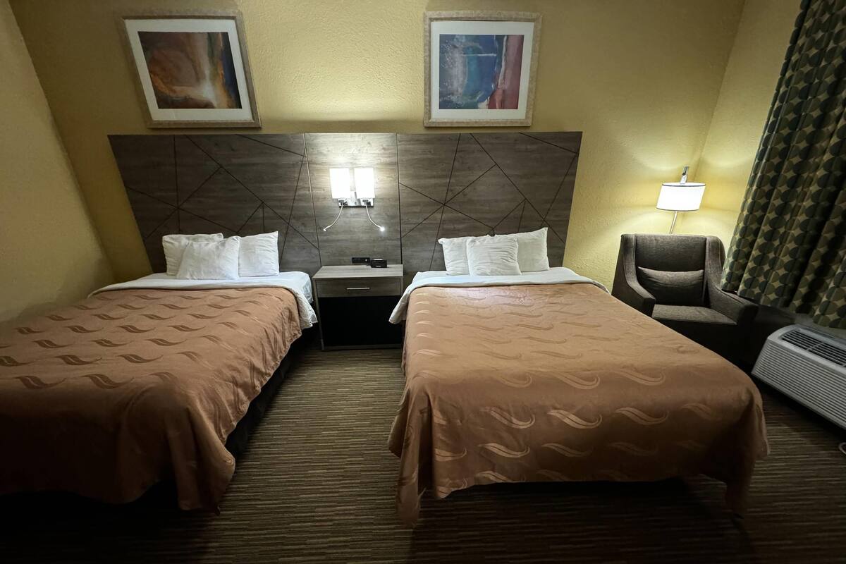 Habitación de hotel con dos camas, una mesita de noche, una silla, una lámpara y dos cuadros en la pared.
