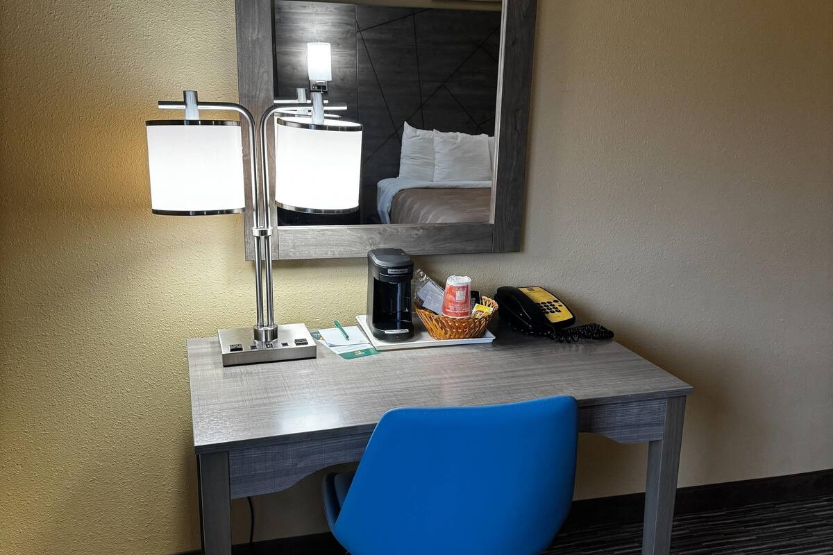 Escritorio de habitación de hotel con lámpara, cafetera, cesta de artículos de aseo, teléfono y una silla azul.