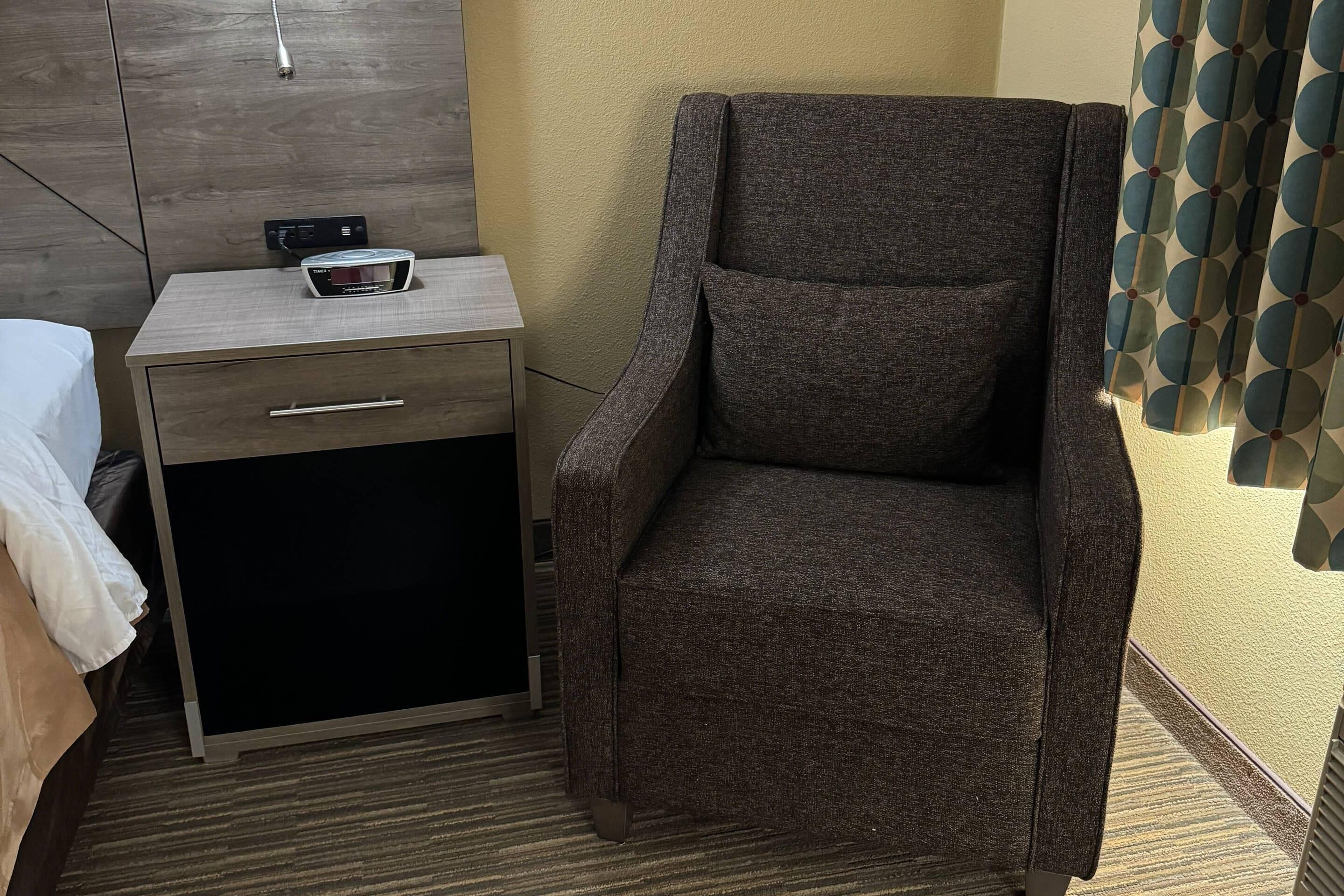 A small room with a bed, a nightstand with a power outlet, a chair, and a patterned curtain.