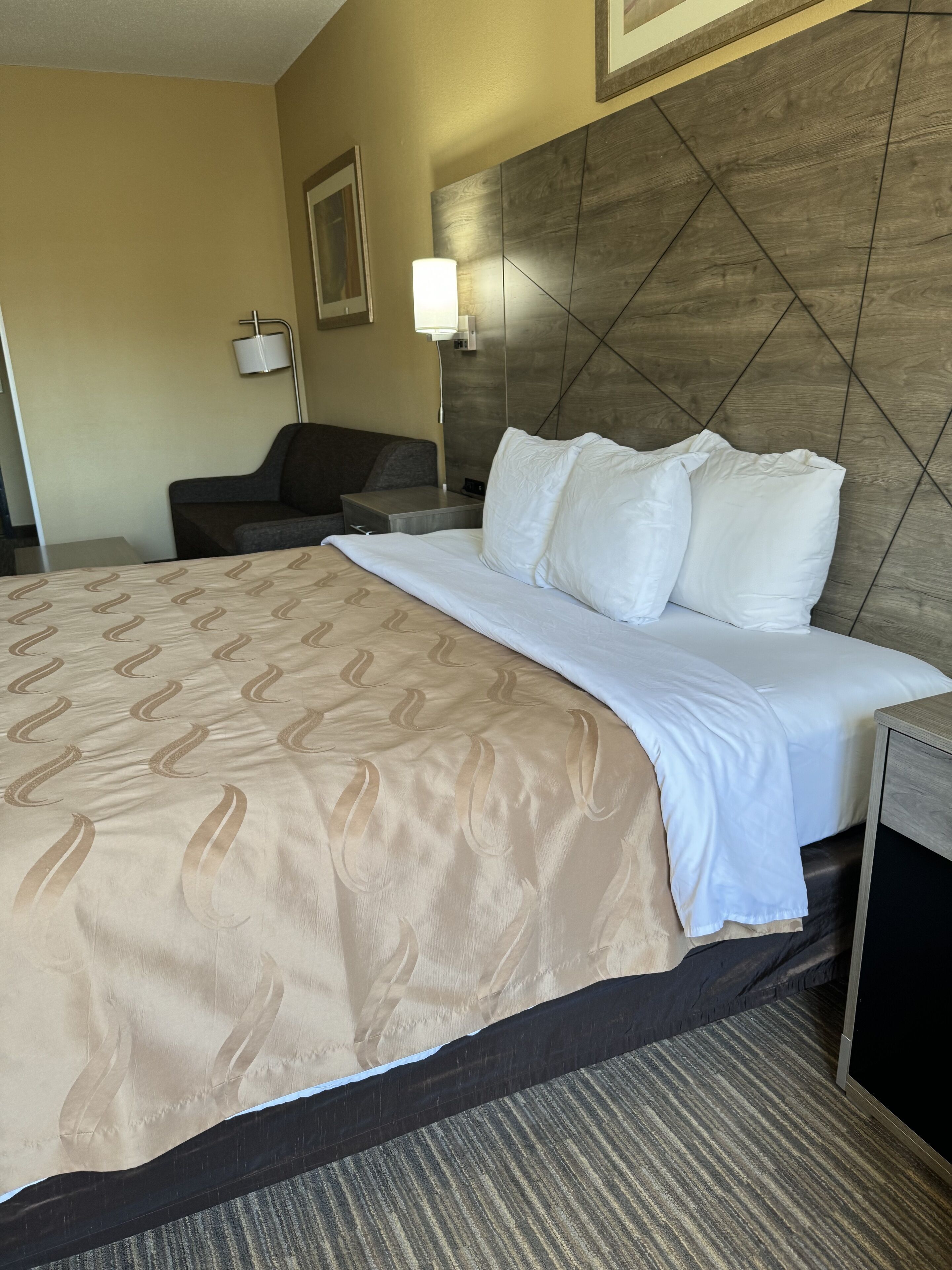 A hotel room with a large bed, a headboard with a geometric pattern, a bedside table, a lamp, and a small couch.