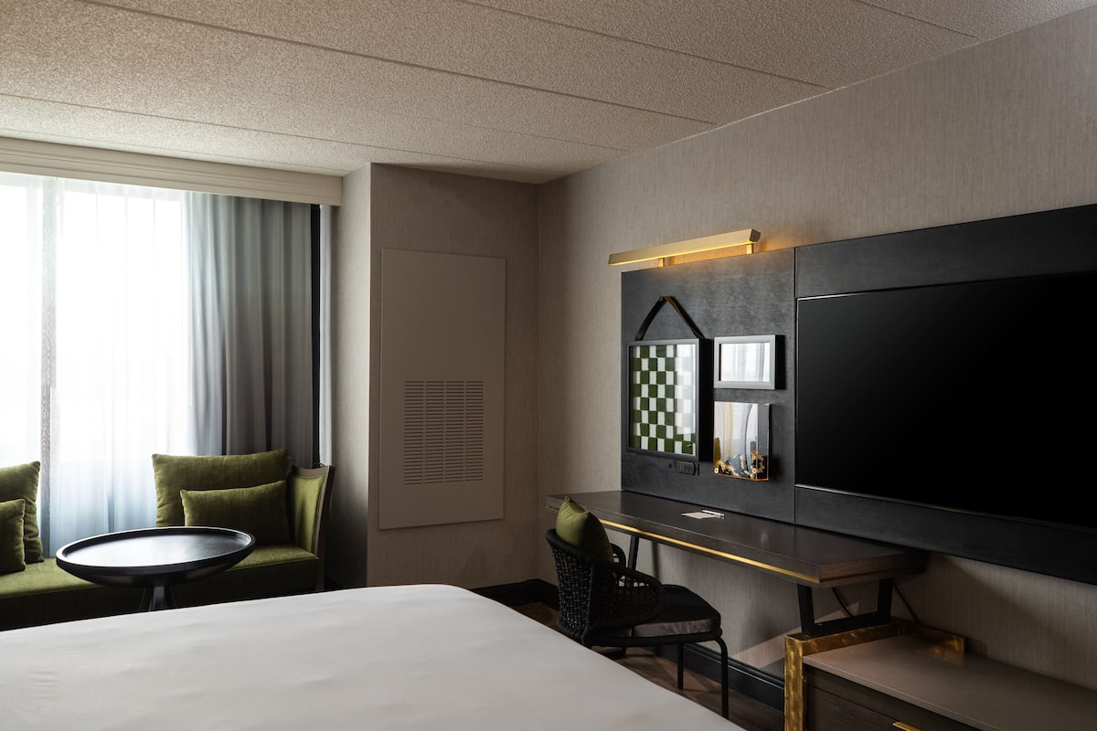 Una camera d'hotel con un letto, una scrivania, una sedia, un tavolino, una televisione e una finestra con tende.