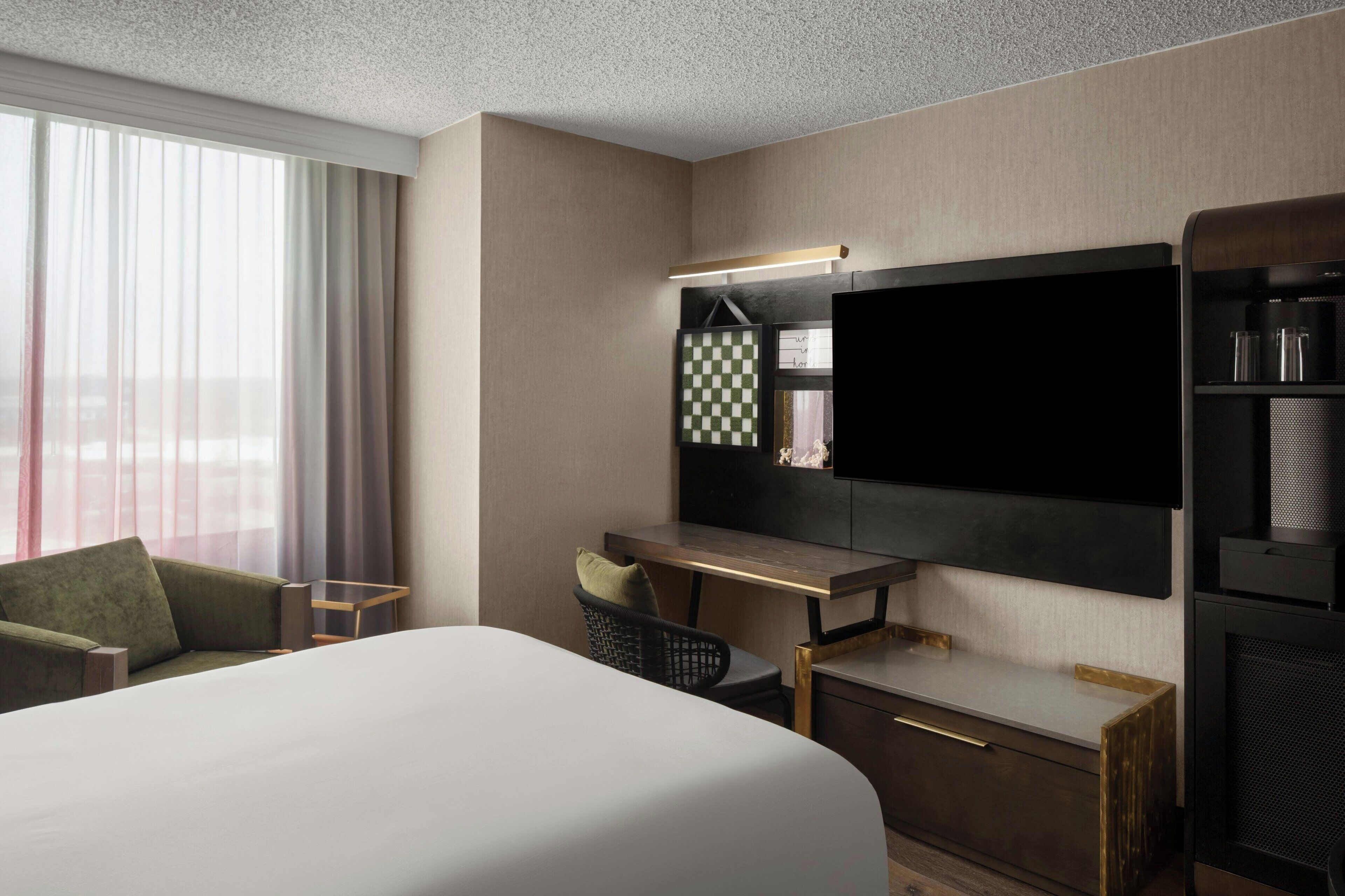 A hotel room with a large bed, a desk with a chair, a flat-screen TV, a minibar, and a large window with curtains.