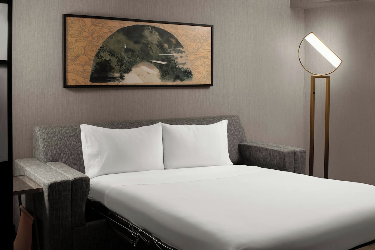 Un letto rifatto con lenzuola bianche, una testiera grigia rivestita in tessuto e un quadro astratto incorniciato sopra.