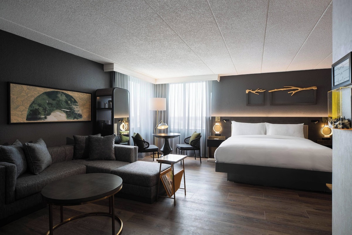 Una moderna camera d'hotel con un divano componibile, un tavolino rotondo, un letto e una scrivania con una lampada.