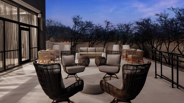 Un espace extérieur aménagé avec des chaises et un canapé en osier, entouré d’arbres et bordé par la façade d’un bâtiment.