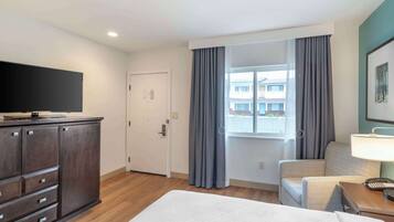 Quarto de hotel com cama, escrivaninha com luminária, cadeira, televisão sobre um móvel e janela com cortinas.