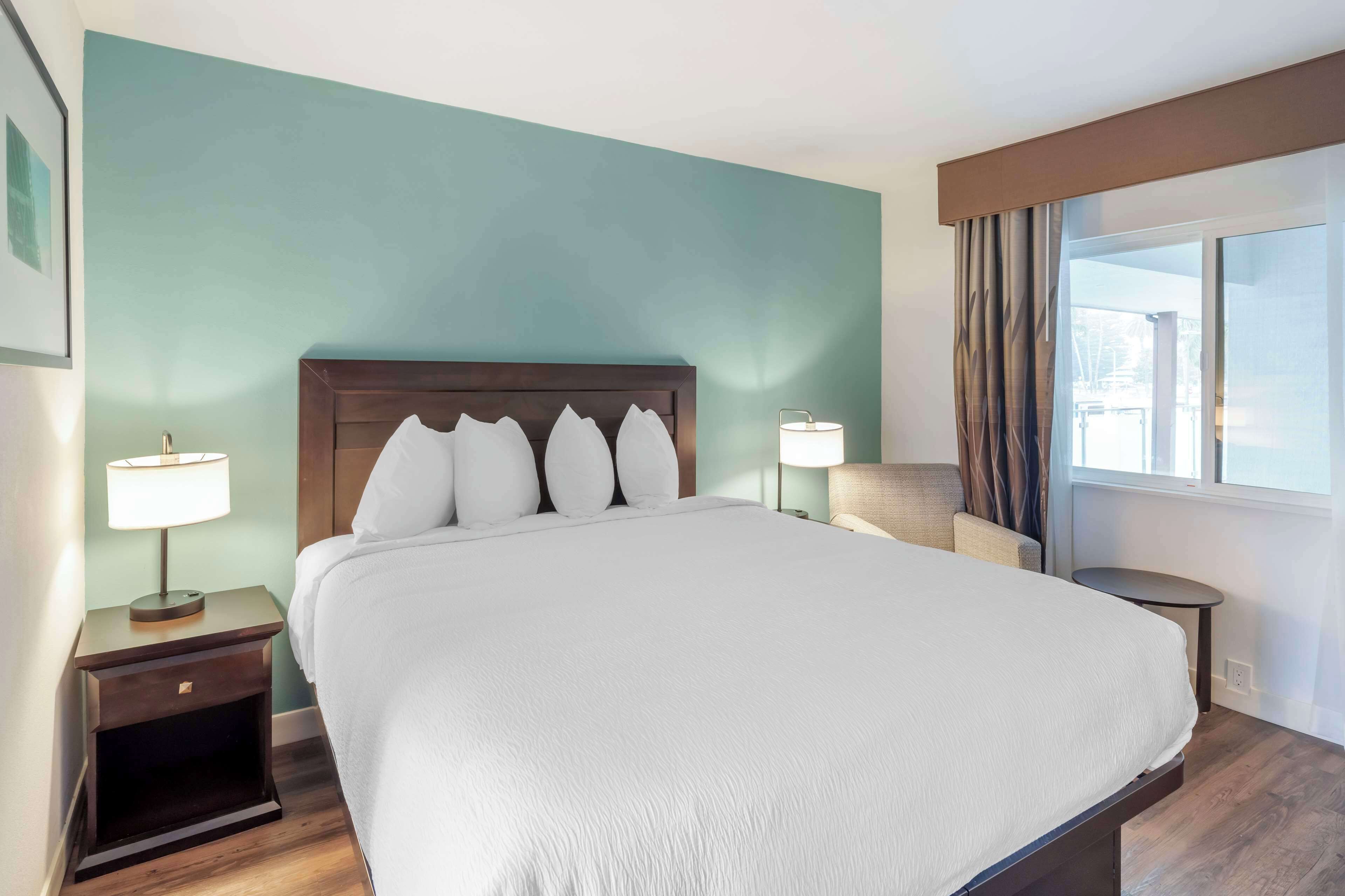 Ein Hotelzimmer mit einem großen Bett, einem Nachttisch mit Lampe, einem Sessel und Blick ins Freie durch ein Fenster.