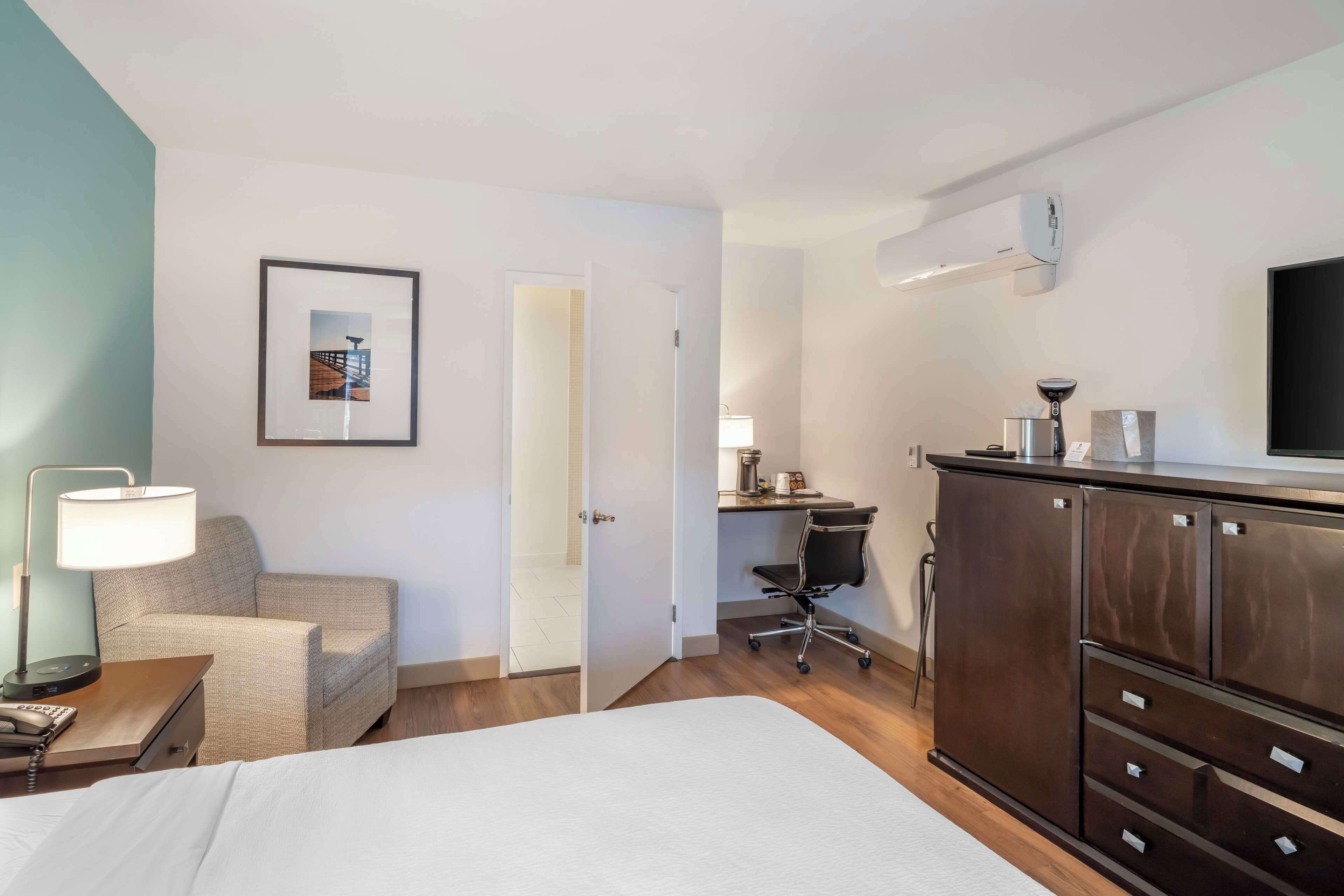 A hotel room with a bed, a desk with a chair, a wardrobe, a bedside table with a lamp, and a framed picture on the wall.