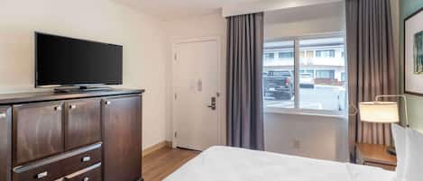 Ein Hotelzimmer mit Bett, einem Flachbildfernseher auf einer Kommode, einem Schreibtisch mit Lampe und Blick auf den Parkplatz durch das Fenster.