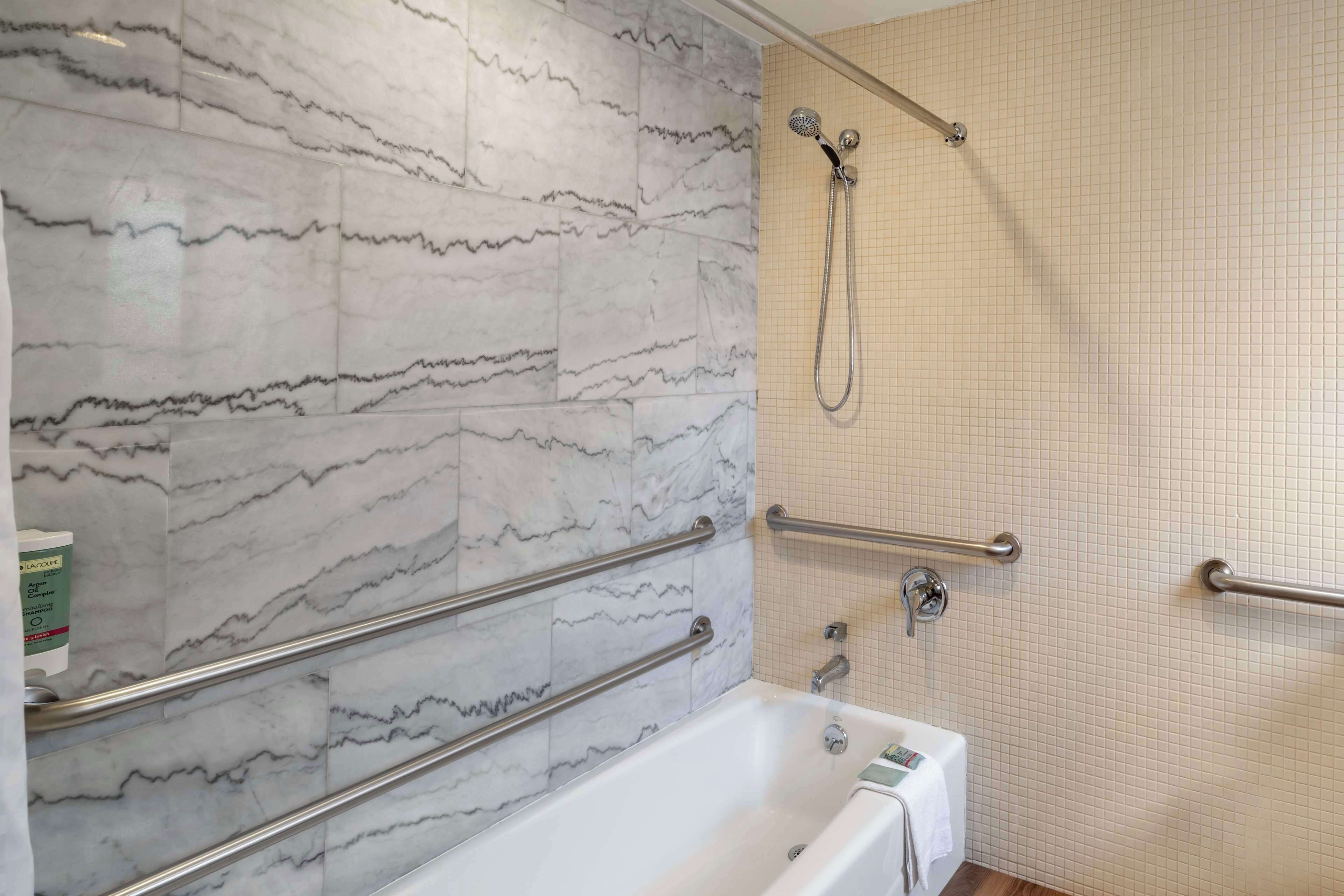 A bathroom with marble walls, a bathtub, a shower with a handheld showerhead, and grab bars.