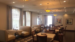 Comedor de hotel con varias mesas y sillas, ventanas amplias con cortinas e iluminación en el techo.