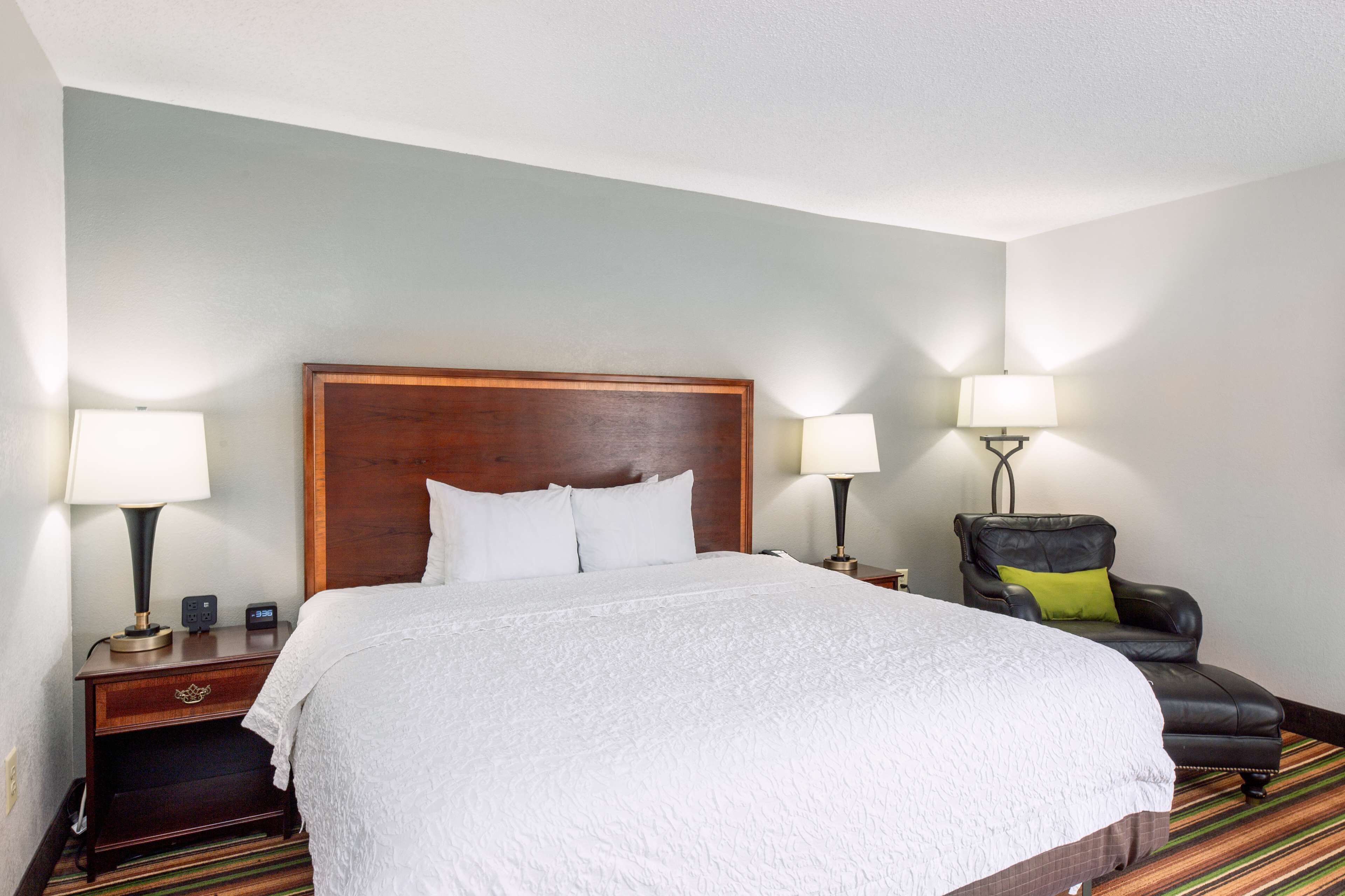 A hotel room with a large bed, two bedside tables with lamps, a chair, and a wooden headboard.
