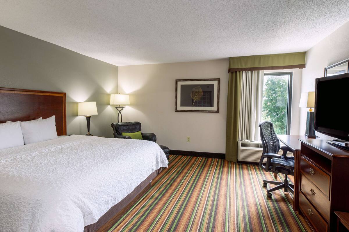 A hotel room with a large bed, a desk with a chair, a television, and a window with curtains.