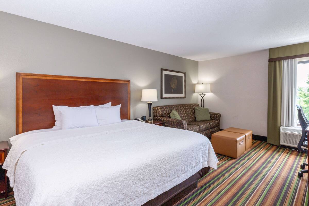 A hotel room with a large bed, a sofa, a desk, a lamp, and a window with curtains.