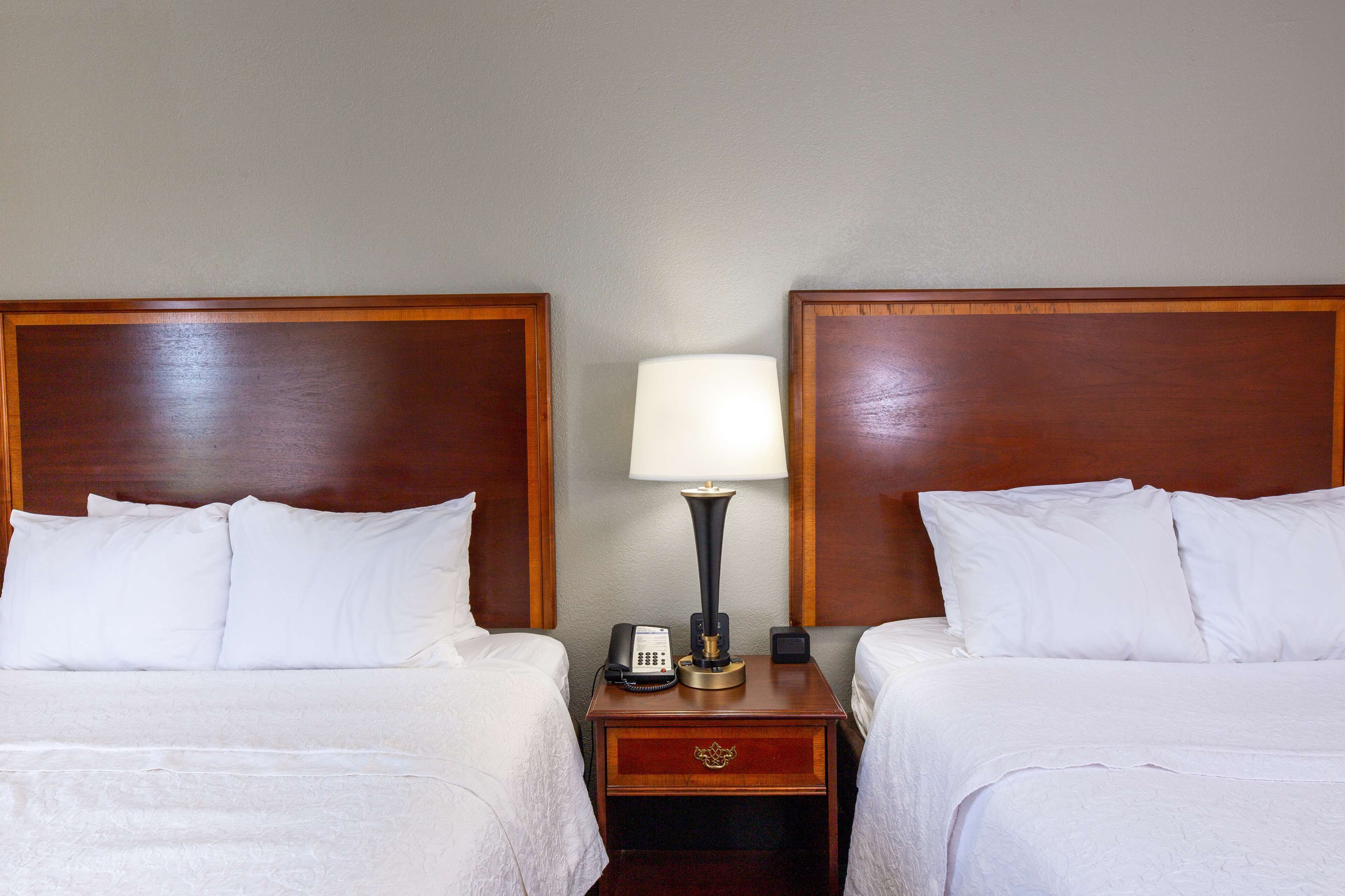 Habitación de hotel con dos camas individuales, cada una con ropa de cama blanca y cabecero de madera. Hay una mesita de noche entre las camas con una lámpara, un teléfono y un cajón pequeño.