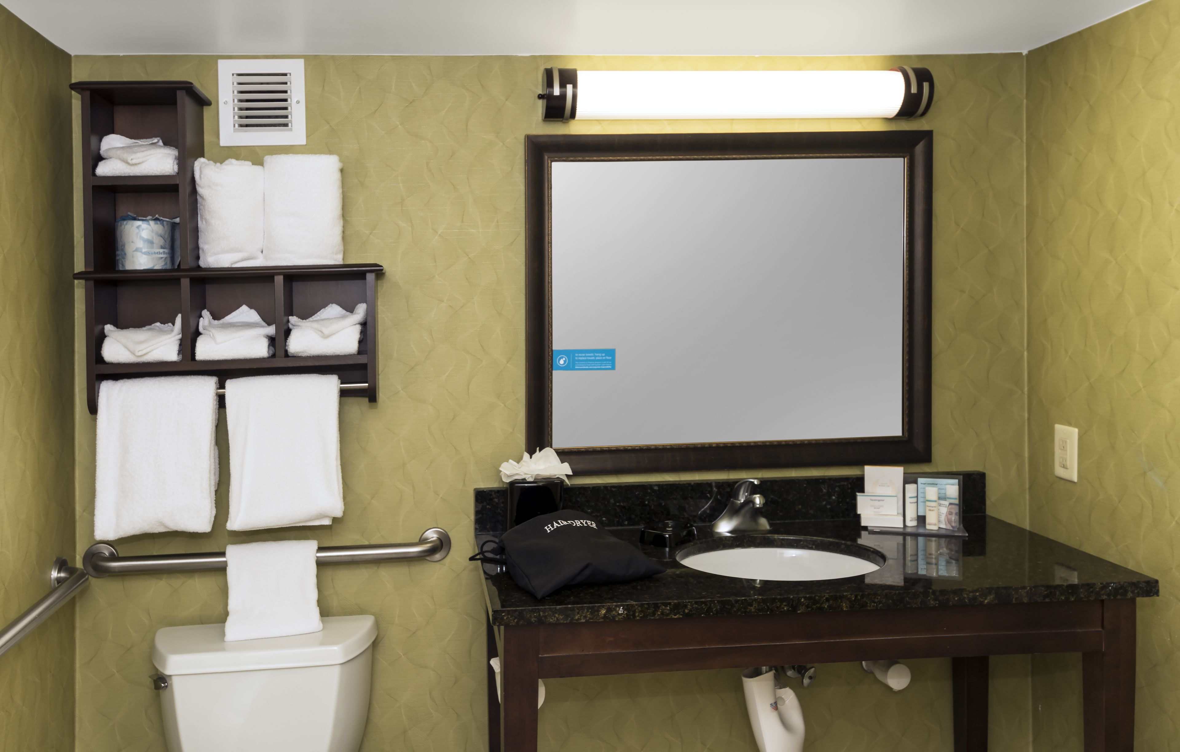 Une salle de bain avec des toilettes, un lavabo et un miroir. Des serviettes sont posées sur une étagère et un sèche-mains est fixé au-dessus du miroir.