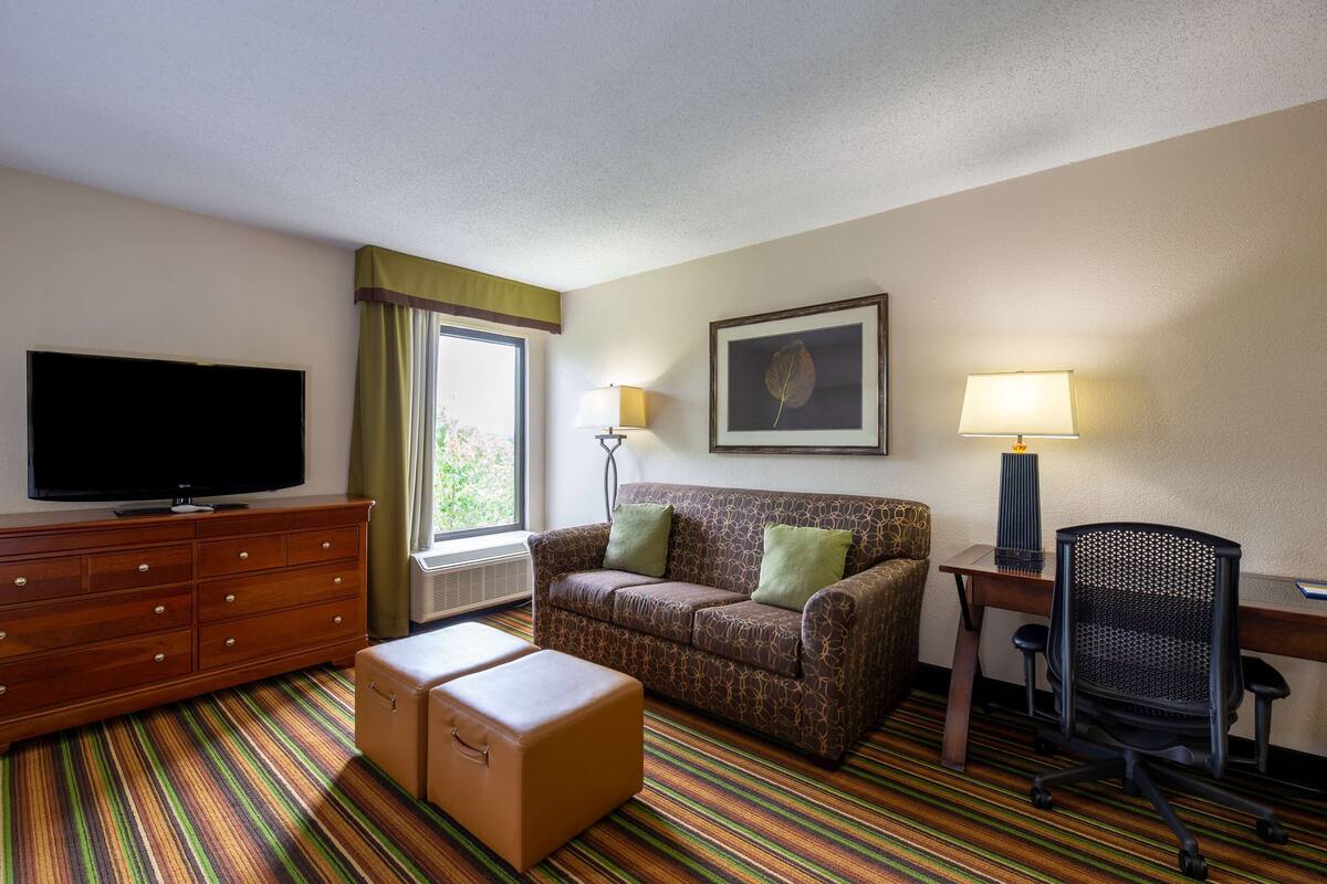 A hotel room with a flat-screen TV, a sofa, a desk with a chair, a lamp, and a window with curtains.
