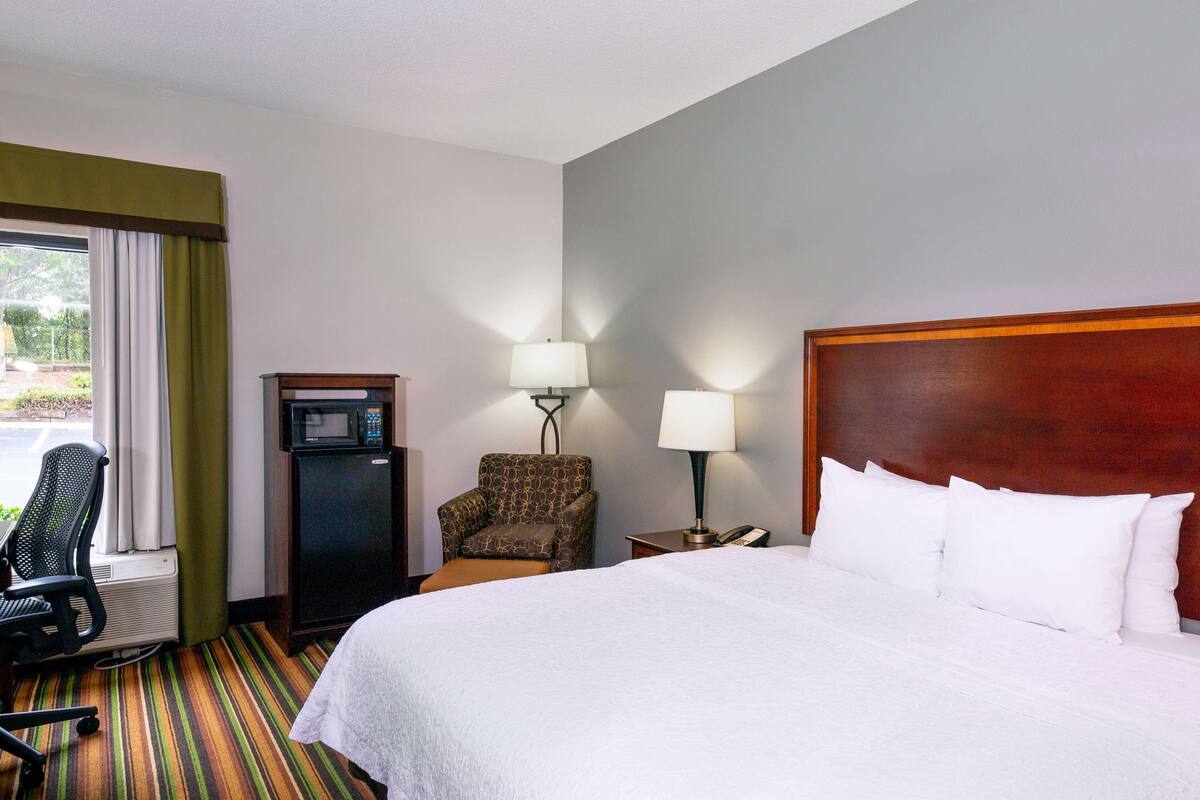 A hotel room with a large bed, a chair, a desk with a lamp, and a microwave.
