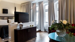 A modern hotel room with a bed, a dark wooden dresser, a flat-screen TV, a glass coffee table with a vase of flowers, and large windows with curtains. - Elite Grand Hotel Norrköping (Norrköping)