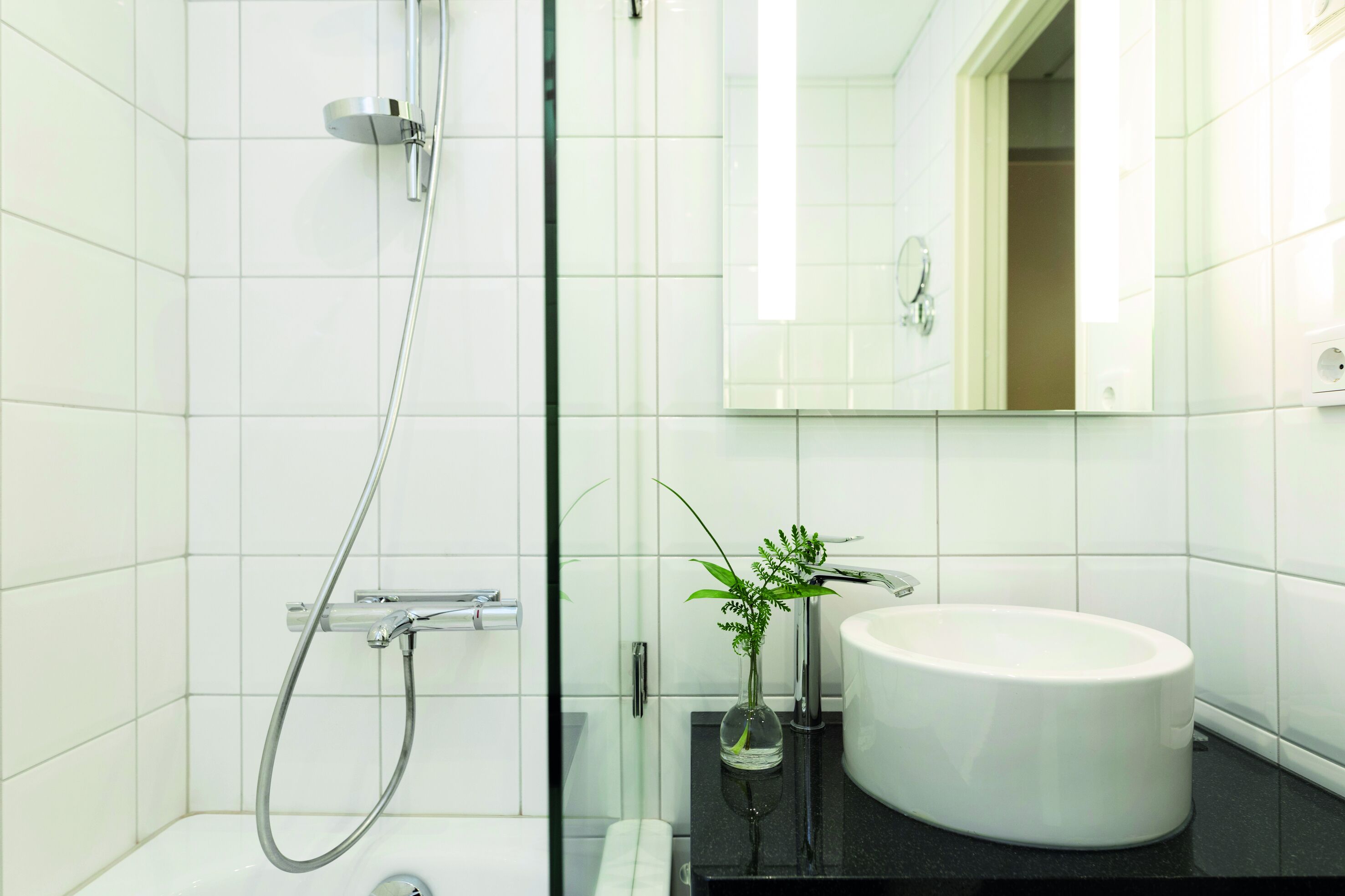 Un baño moderno con azulejos blancos, cabina de ducha de vidrio, lavamanos blanco y espejo sobre el lavamanos.