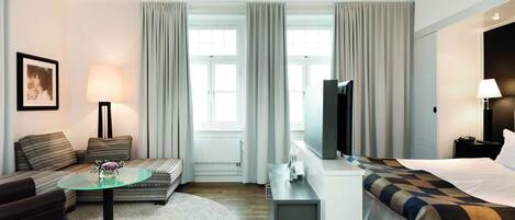 Habitación de hotel con sofá, una mesita de vidrio, cama con colcha estampada, televisión y escritorio con lámpara.