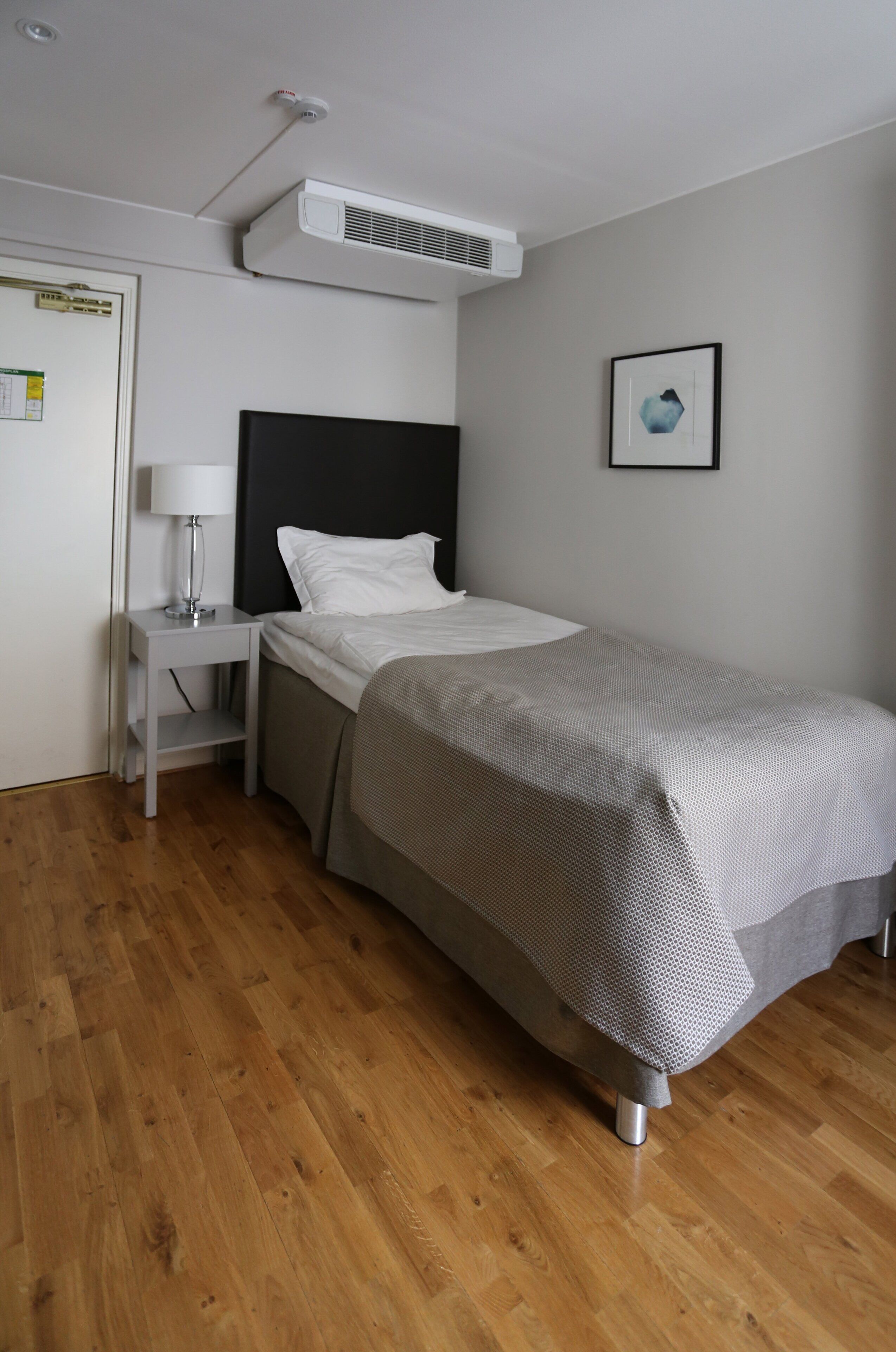 A single bed with a grey and white patterned bedspread, a white nightstand with a lamp, and a wall-mounted air conditioner.