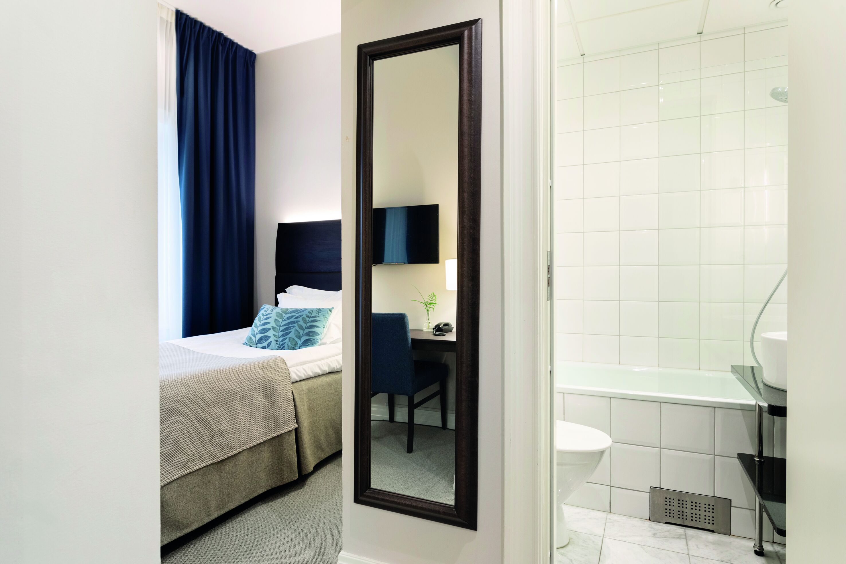 A hotel room with a bed, a desk with a chair, a mirror, and a bathroom with a bathtub and sink.
