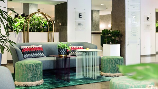 A modern hotel lobby with a grey sofa, green ottomans, a round metal table, and potted plants. - Elite Grand Hotel Norrköping (Norrköping)