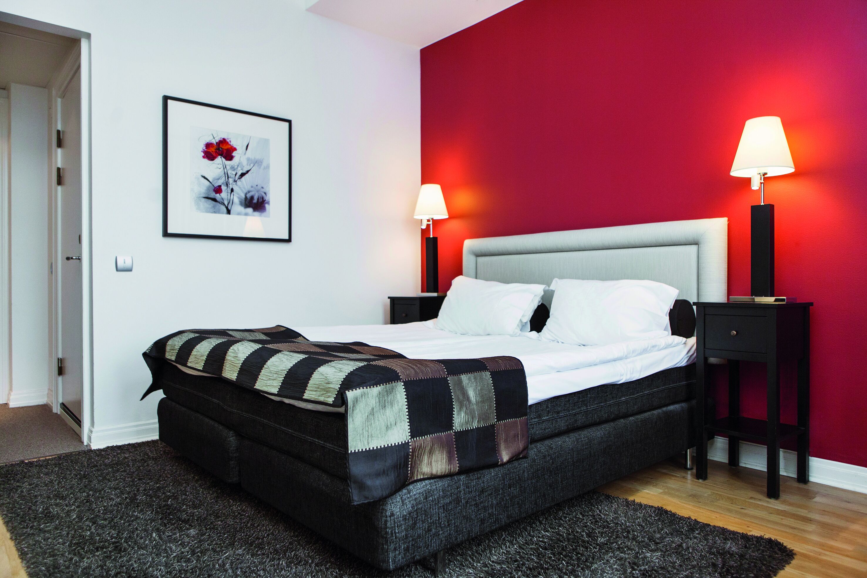 A modern bedroom with a large bed, two bedside tables with lamps, a red accent wall, and a framed artwork above the bed.