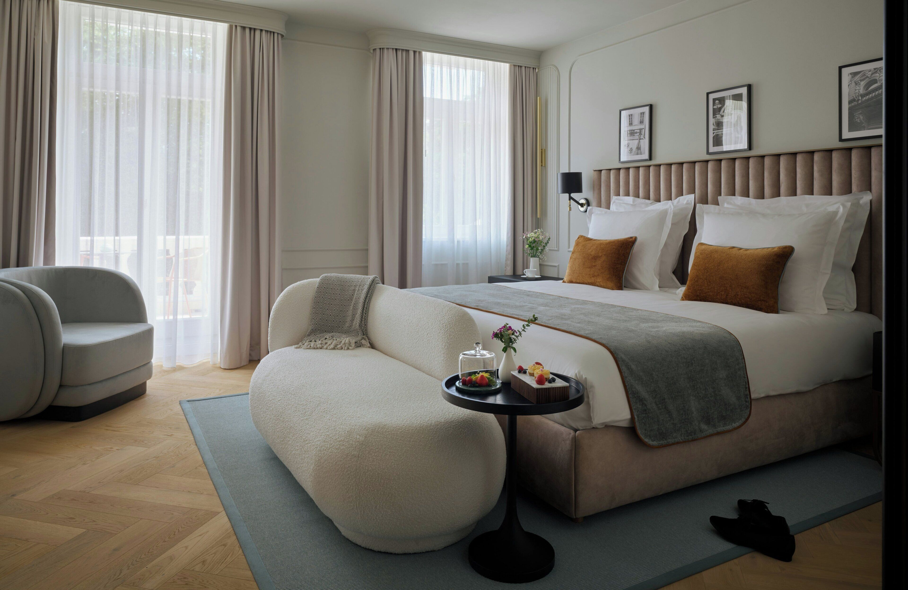 Quarto de hotel moderno com uma cama grande, um sofá, uma mesinha de centro com um vaso e uma bandeja de frutas, e vista para o exterior por grandes janelas.