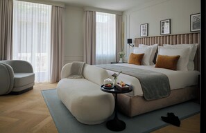 Una habitación de hotel moderna con una cama grande, un sofá, una mesita con un florero y una bandeja de fruta, y vistas al exterior a través de amplios ventanales.