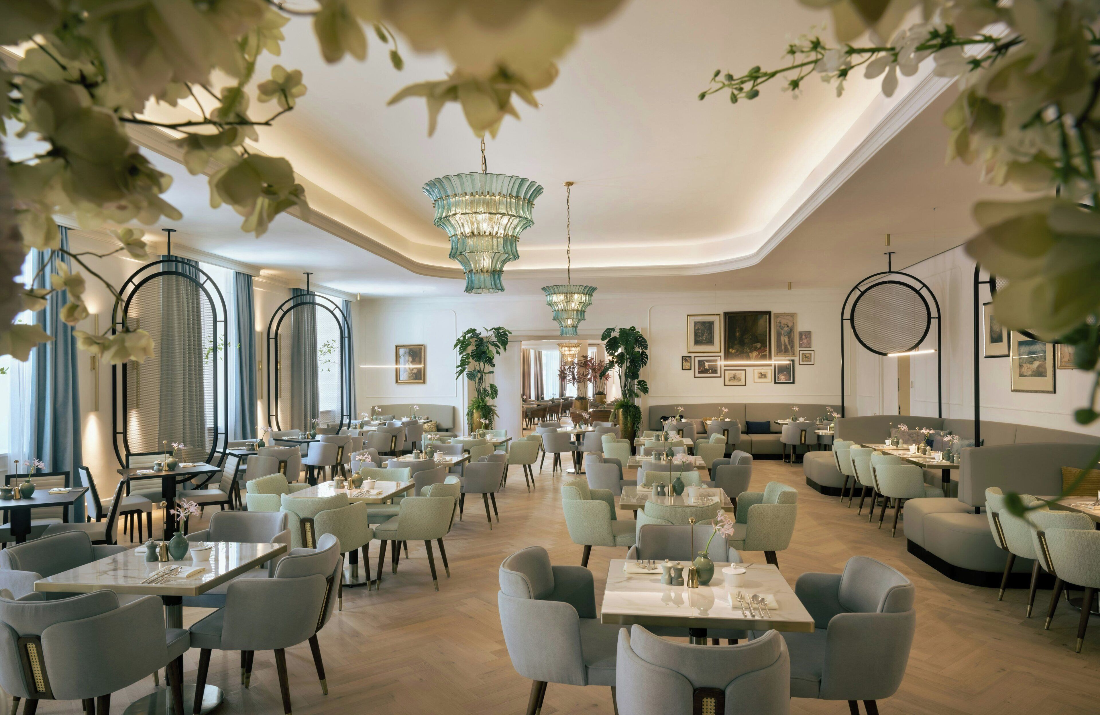 Ein großzügiges Restaurant mit verschiedenen Sitzbereichen, darunter runde Tische und gemütliche Sitzecken, sowie Pflanzen und Kunstwerke an den Wänden.