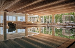 Um espaço amplo e interno com uma piscina espelhada, teto de madeira e grandes janelas que oferecem vista para o verde.