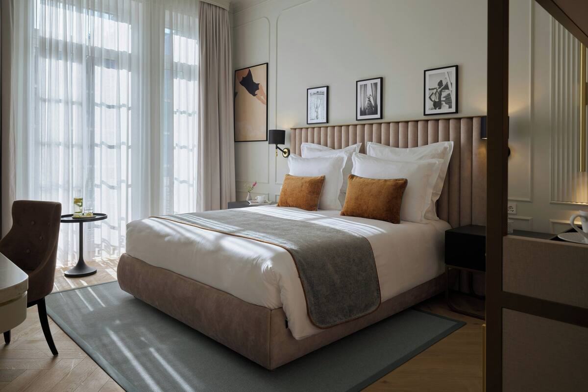 A hotel room with a large bed, a bedside table, a chair, and a view of the outdoors through sheer curtains.