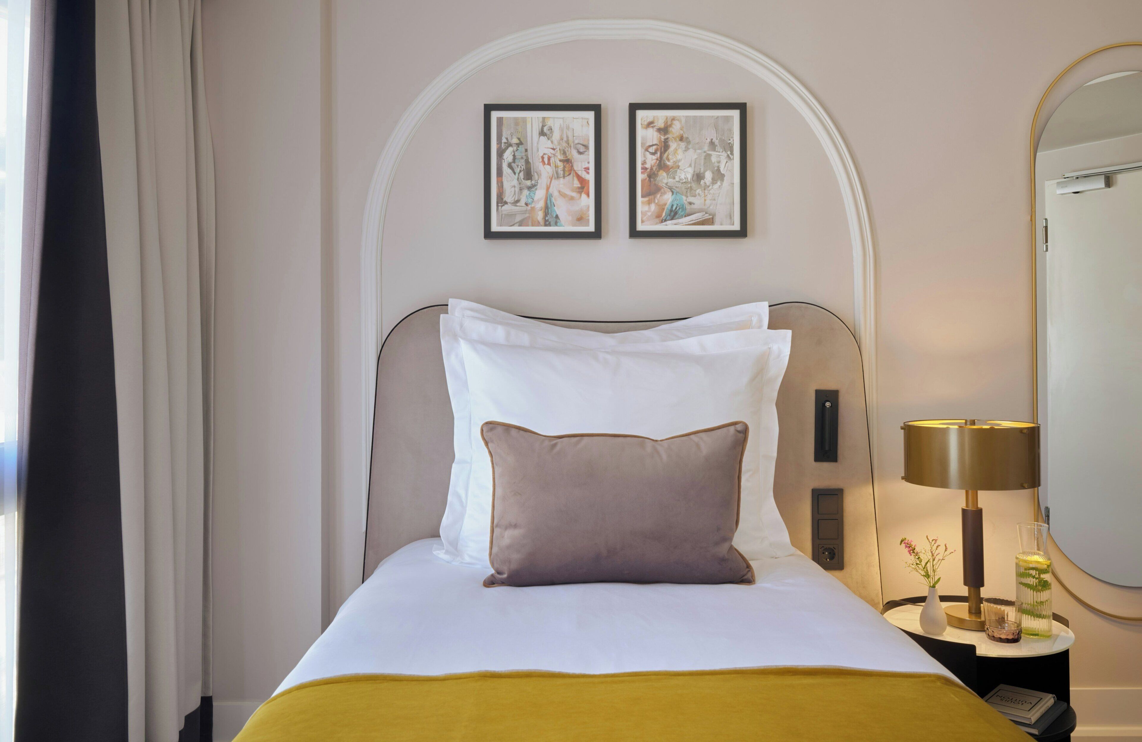 Un lit bien fait, agrémenté d’oreillers blancs et bruns, une table de chevet avec une lampe et un vase, et deux œuvres d’art encadrées accrochées au mur.