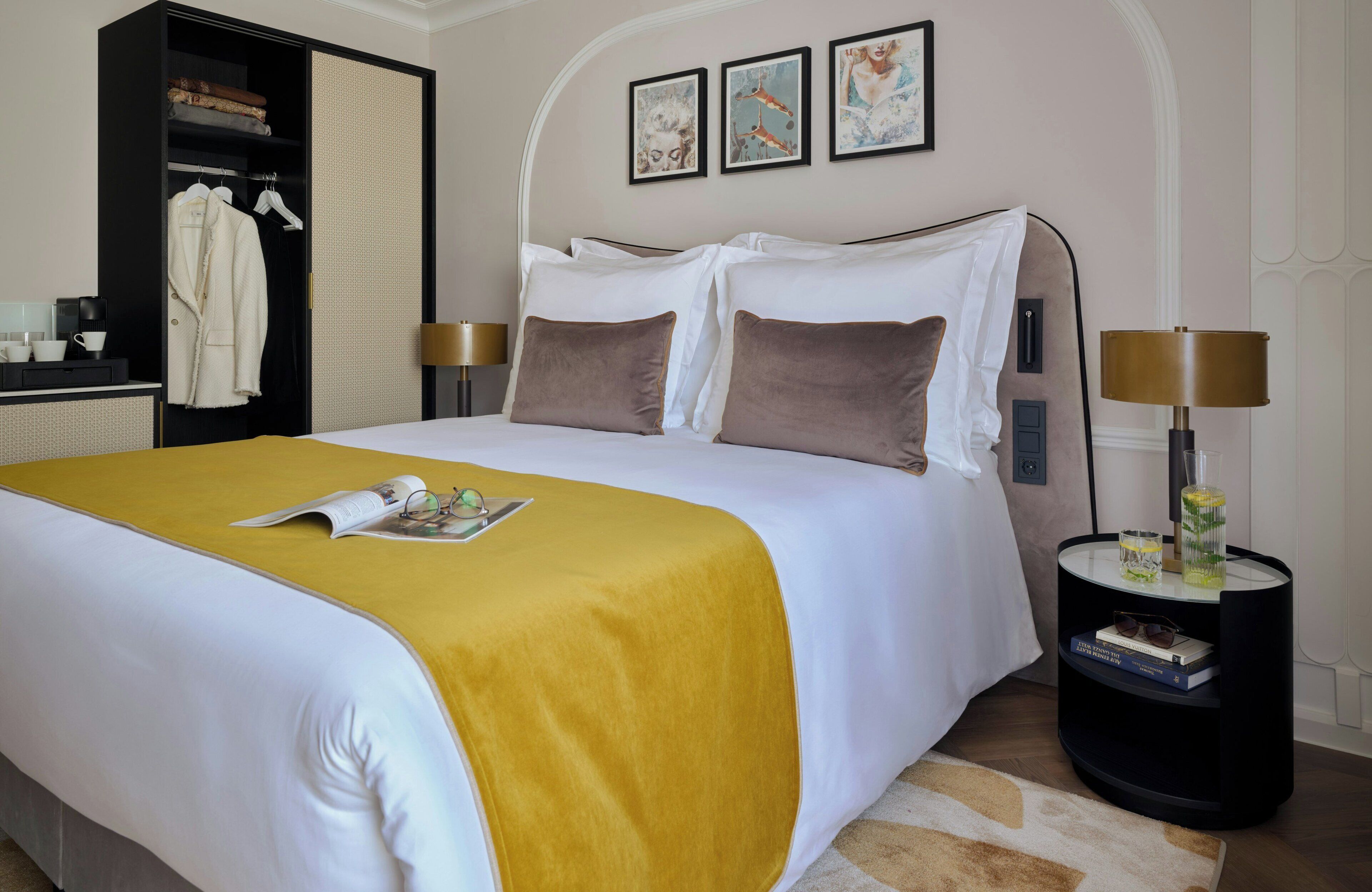 A neatly made bed with a yellow bedspread, a bedside table with a lamp, and a wardrobe with clothes hanging inside.