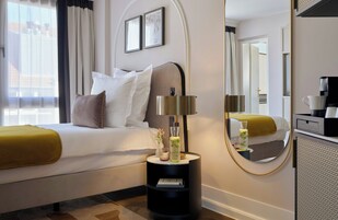 Quarto de hotel com cama, mesa de cabeceira, luminária, espelho e cafeteira.