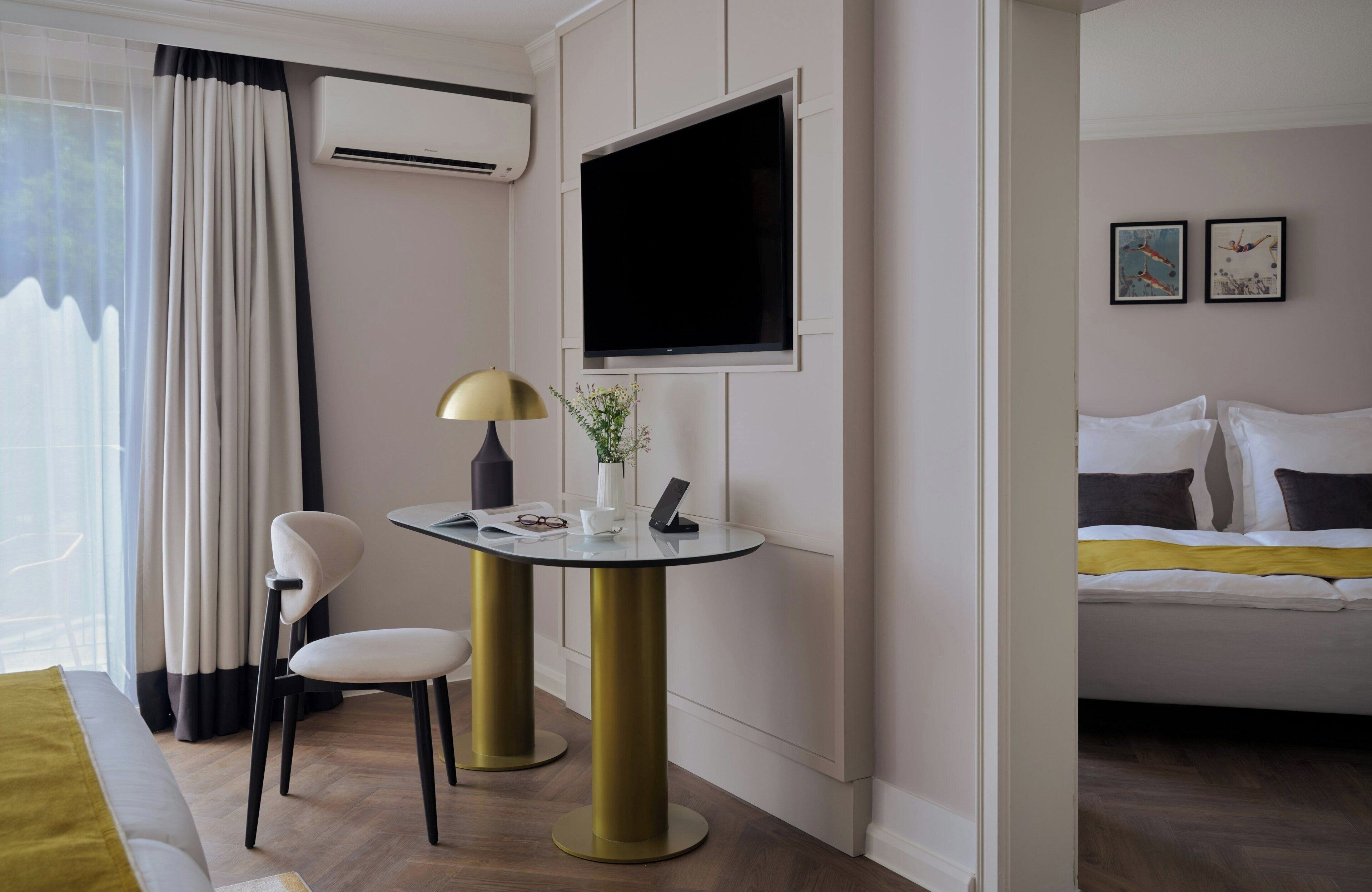 Een hotelkamer met een bed, een aan de muur bevestigde televisie, een klein tafeltje met twee stoelen, een lamp en een raam met gordijnen.
