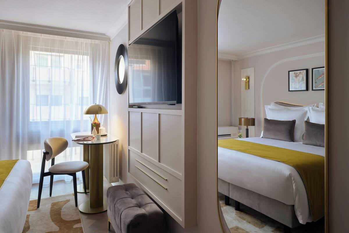 A hotel room with a bed, a desk with a lamp, a chair, a round table, and a television.