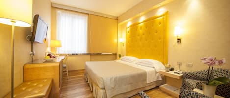 A hotel room with a bed, a desk with a chair, a small table with a lamp, a sofa, and a TV mounted on the wall.