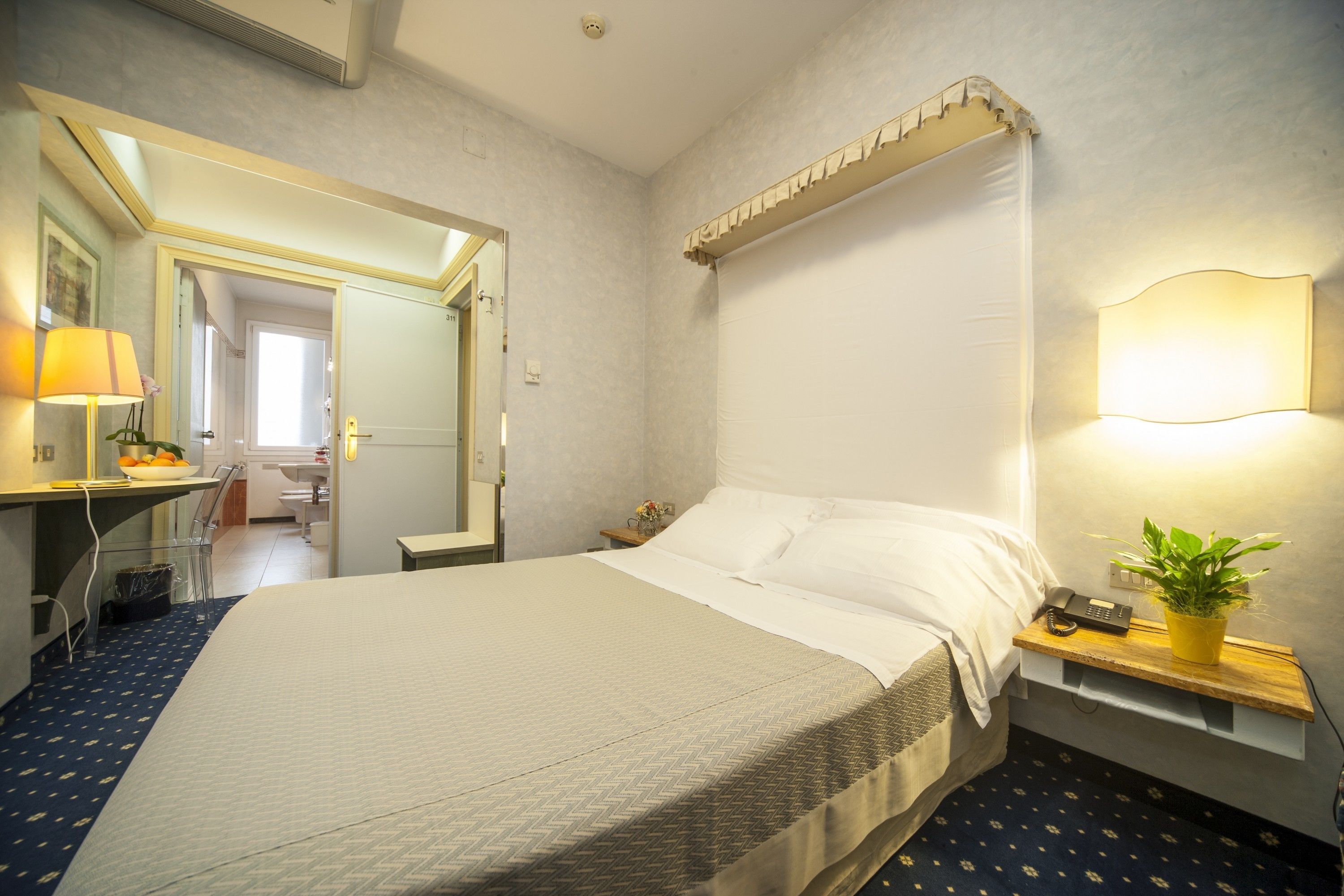 A hotel room with a large bed, a bedside table with a lamp and a plant, a desk with a lamp, and a mirror.