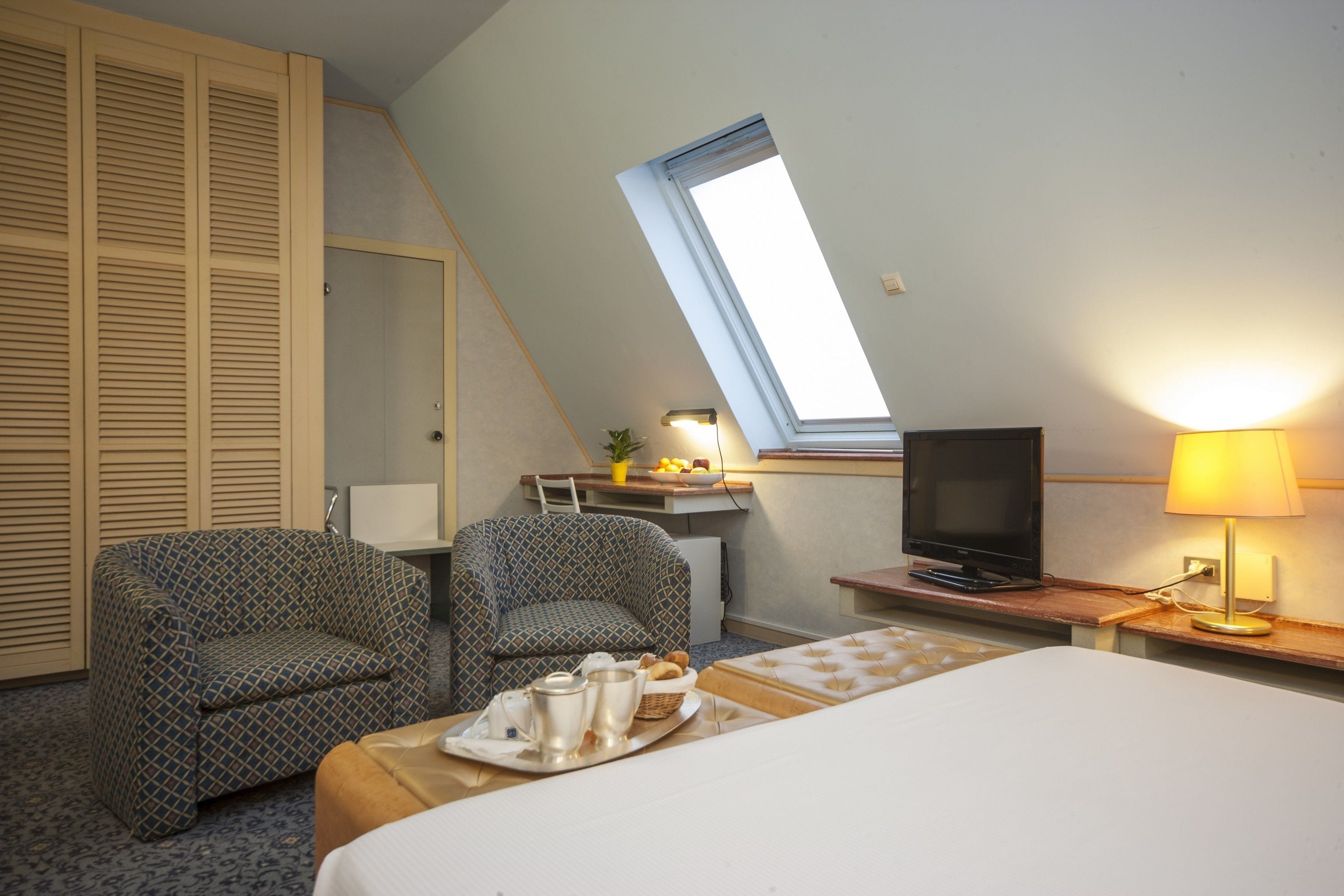 Une chambre d’hôtel avec un lit, deux fauteuils à motifs, un bureau avec une télévision, une lampe et une lucarne.