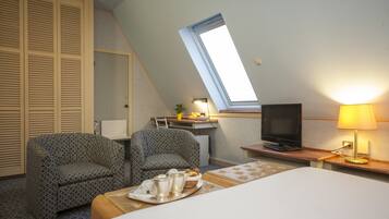 Une chambre d’hôtel avec un lit, deux fauteuils à motifs, un bureau avec une télévision, une lampe et une lucarne.